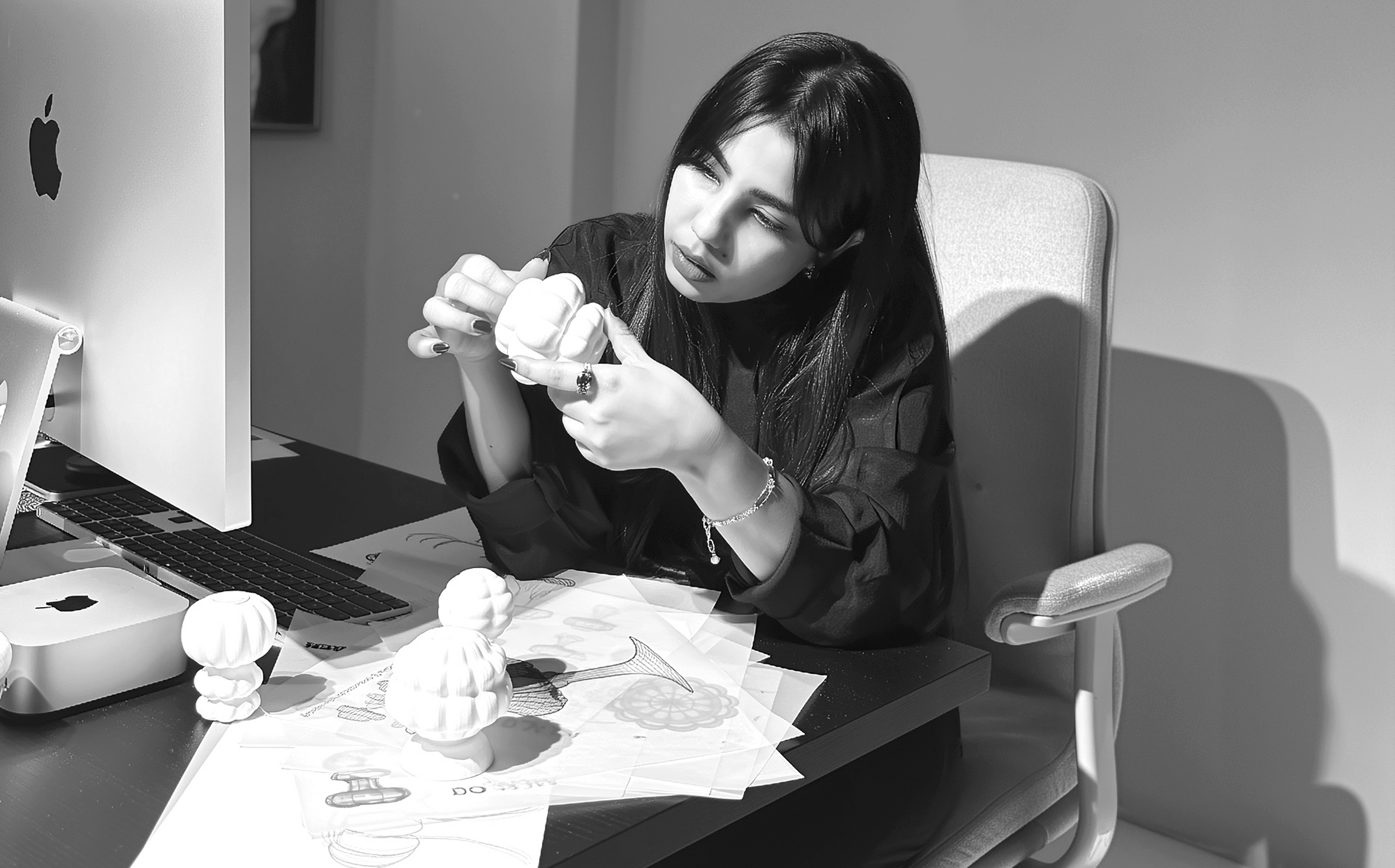Sana Alam, founder of Mugen, studying a lamp prototype at her studio desk surrounded by sketches and design sheets.